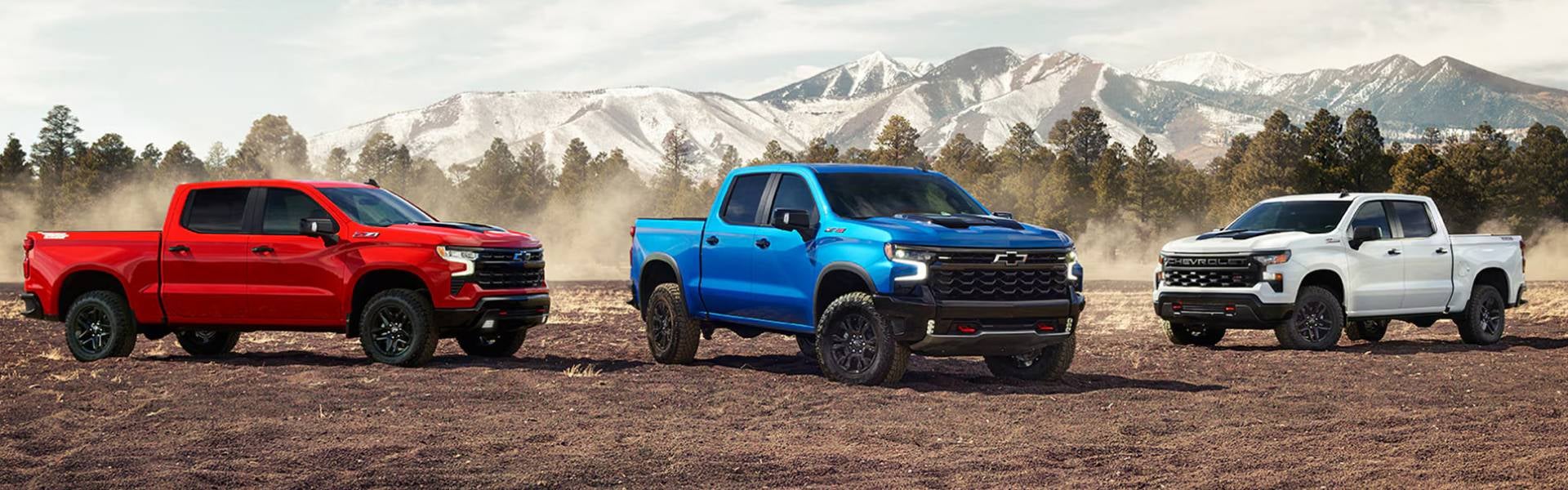 New Chevrolet trucks lined up in the dusty ranch infront of a snowy mountain and trees.